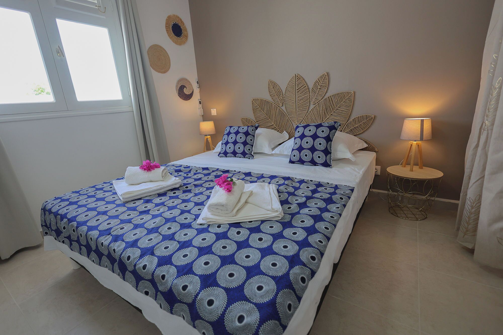 A hotel room with a bed, two bedside lamps, a wall with decorative plates, and a window with curtains.