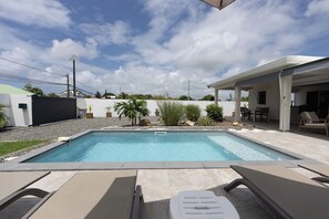 A swimming pool with lounge chairs, a covered patio area, and a view of a residential neighborhood.