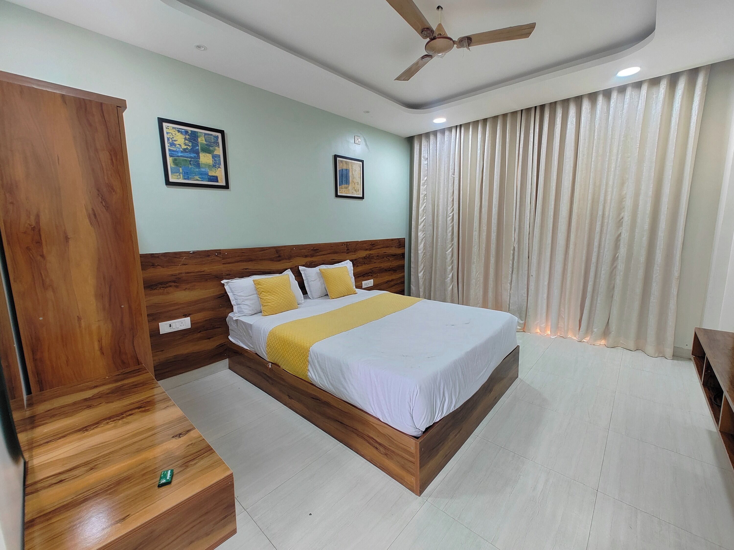 A bedroom with a wooden bed frame, white bedding, and a yellow throw. There is a ceiling fan, a painting on the wall, and a window with curtains.