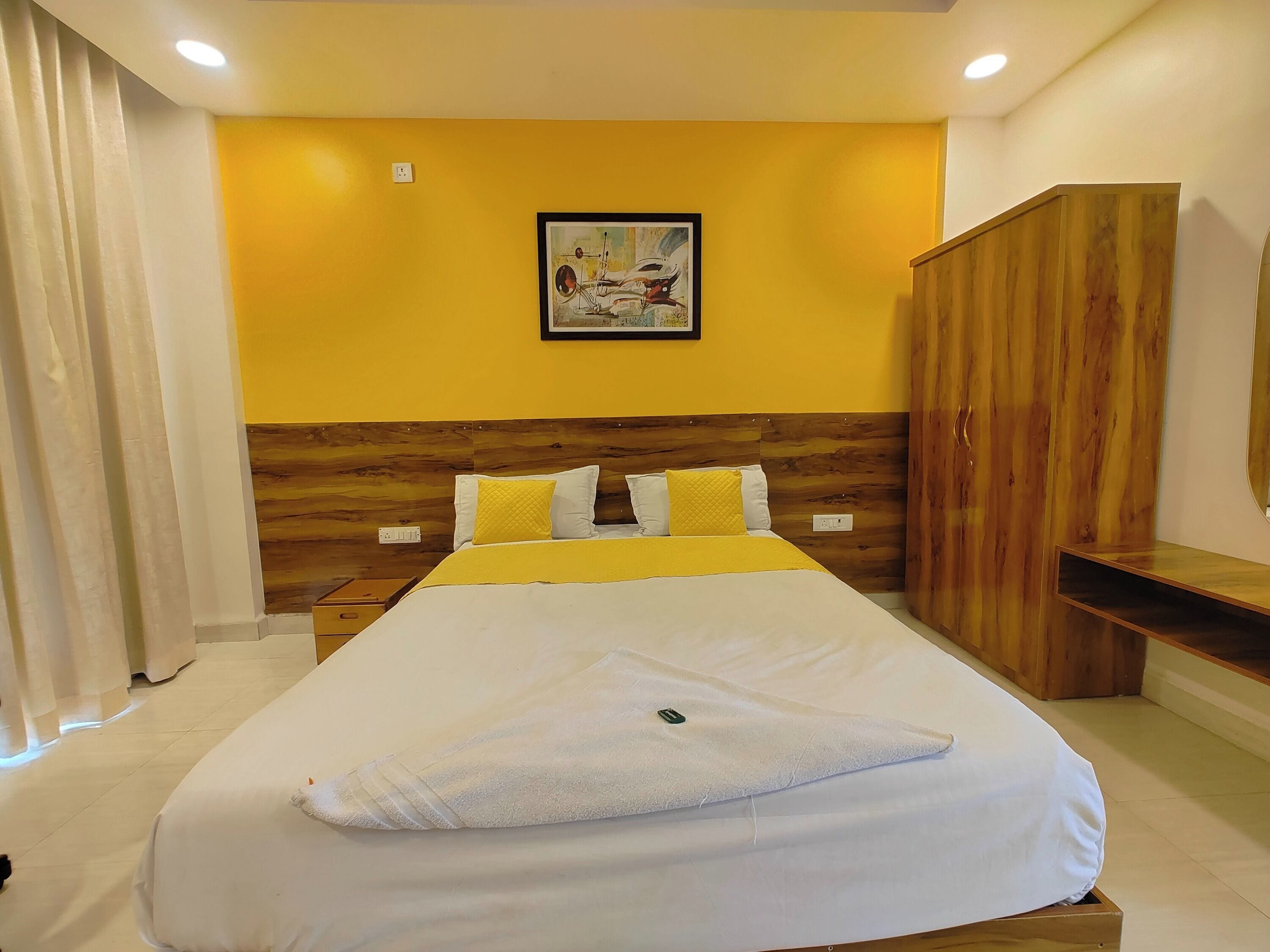 A hotel room with a large bed, a wooden headboard, a yellow accent wall, and a painting above the bed.