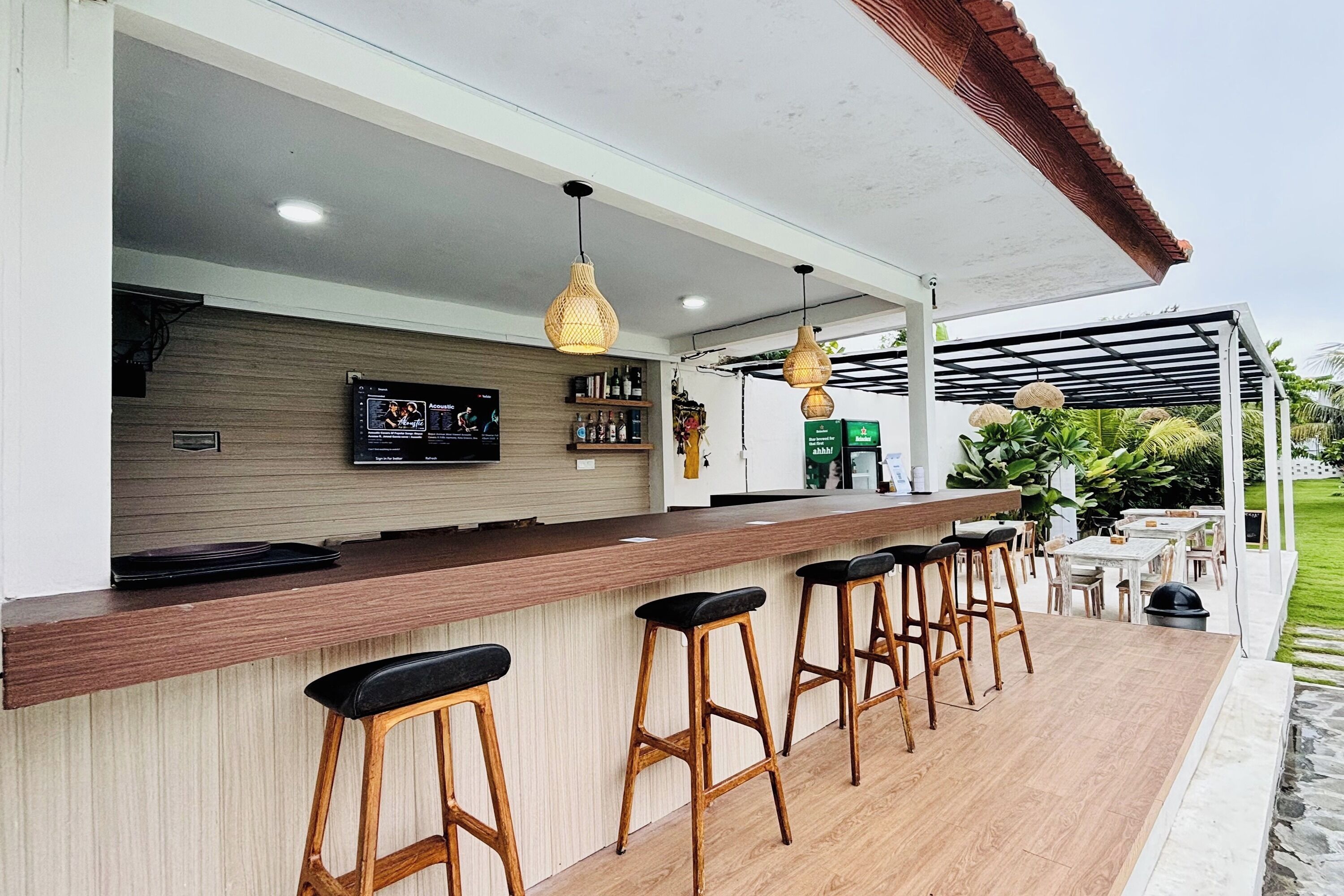 A modern outdoor bar area with a wooden counter, bar stools, hanging wicker lights, and a view of a garden.
