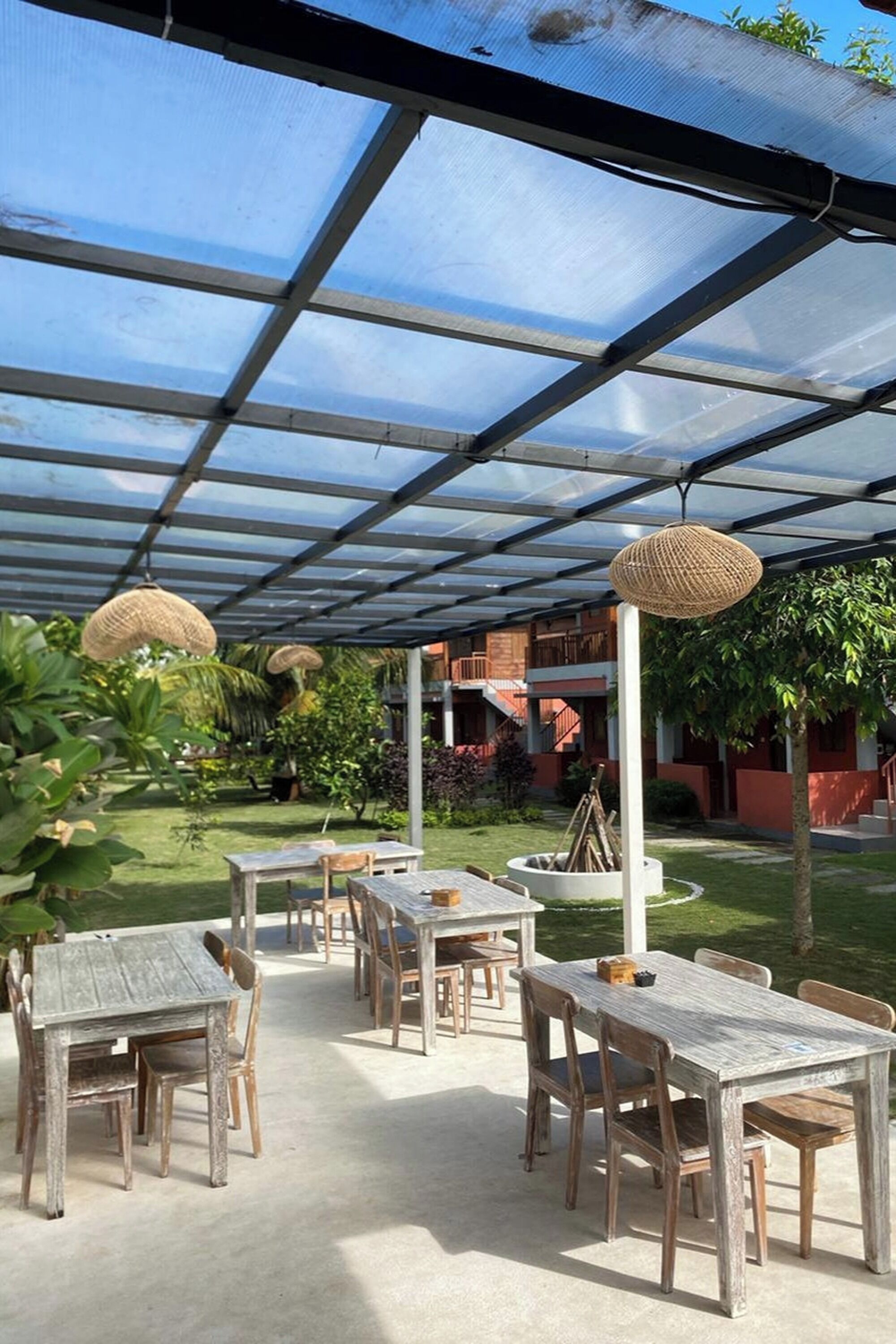 An outdoor patio with wooden tables and chairs, covered by a transparent roof, surrounded by greenery and a building in the background.
