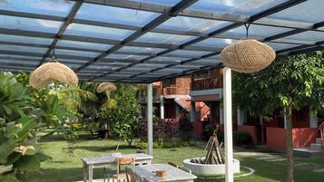 An outdoor patio with wooden tables and chairs, covered by a transparent roof, surrounded by greenery and a building in the background.