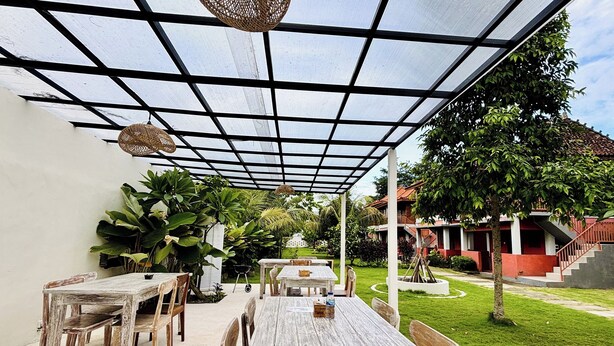 Une terrasse extérieure avec des tables et des chaises en bois, une pergola dotée d’un toit en treillis, et une vue sur un quartier résidentiel avec un immeuble rouge et un escalier.