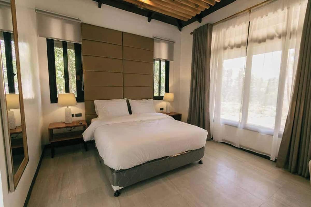 A modern bedroom with a large bed, two bedside tables, and a view of the outdoors through large windows.
