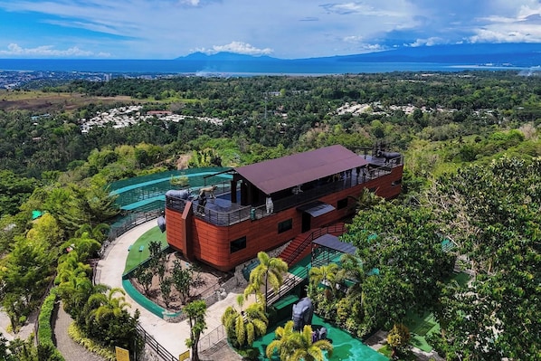 A modern building with a red facade, surrounded by lush greenery and a view of the ocean. - Amaya View (Cagayan de Oro)