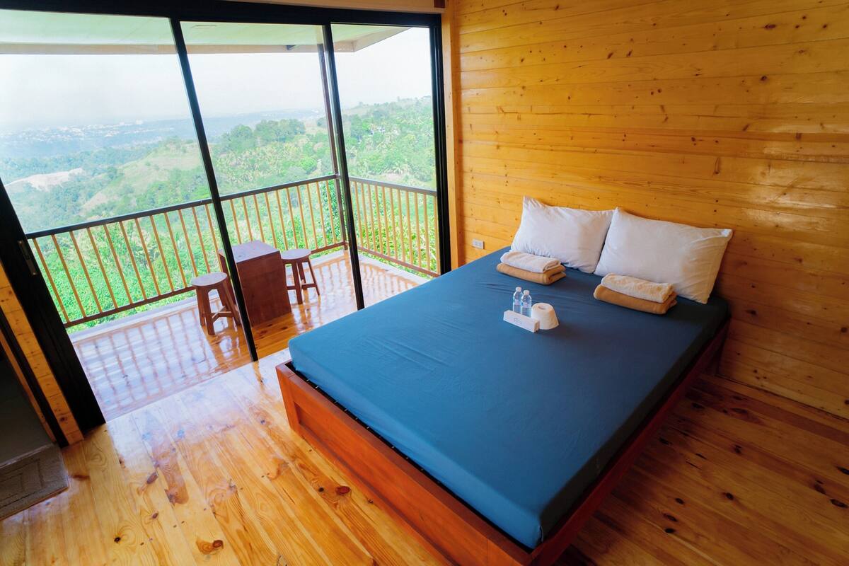 A bedroom with a large bed, wooden walls, and a balcony with a view of the countryside.