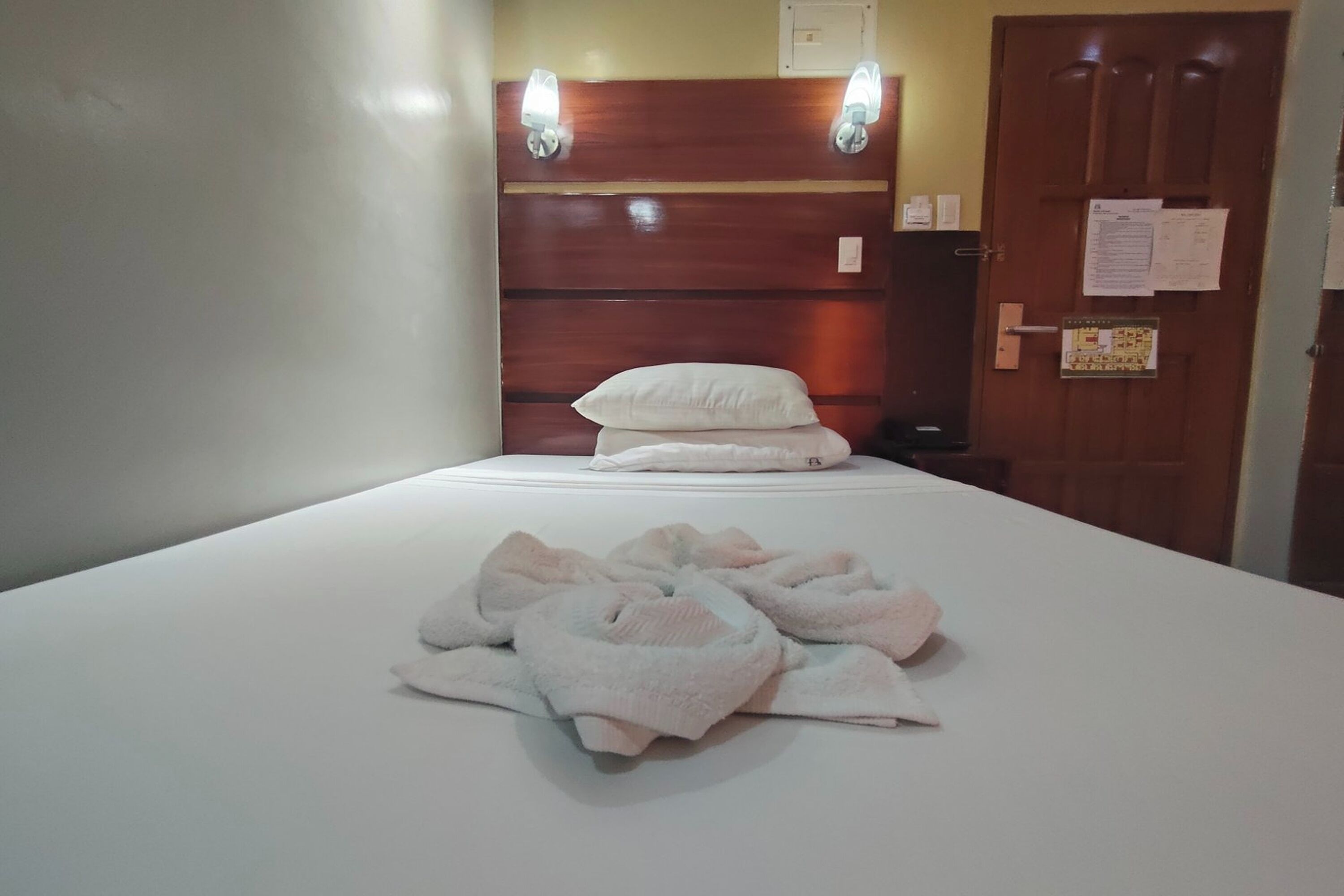 A hotel room with a bed, a wooden headboard, and a door with a notice on it.