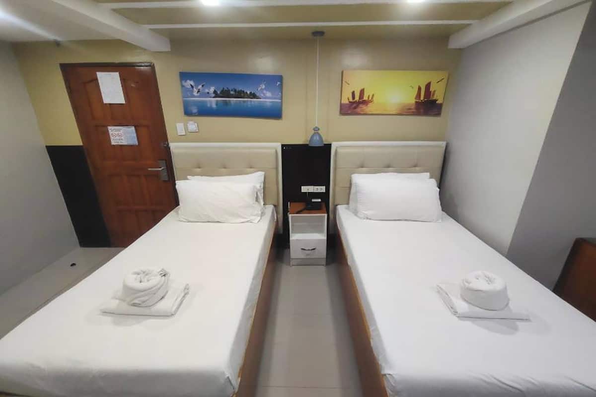 Une chambre d’hôtel avec deux lits simples, chacun agrémenté d’un oreiller blanc et d’une serviette. Il y a une porte sur la gauche, une petite table entre les lits et un mur orné de deux tableaux.