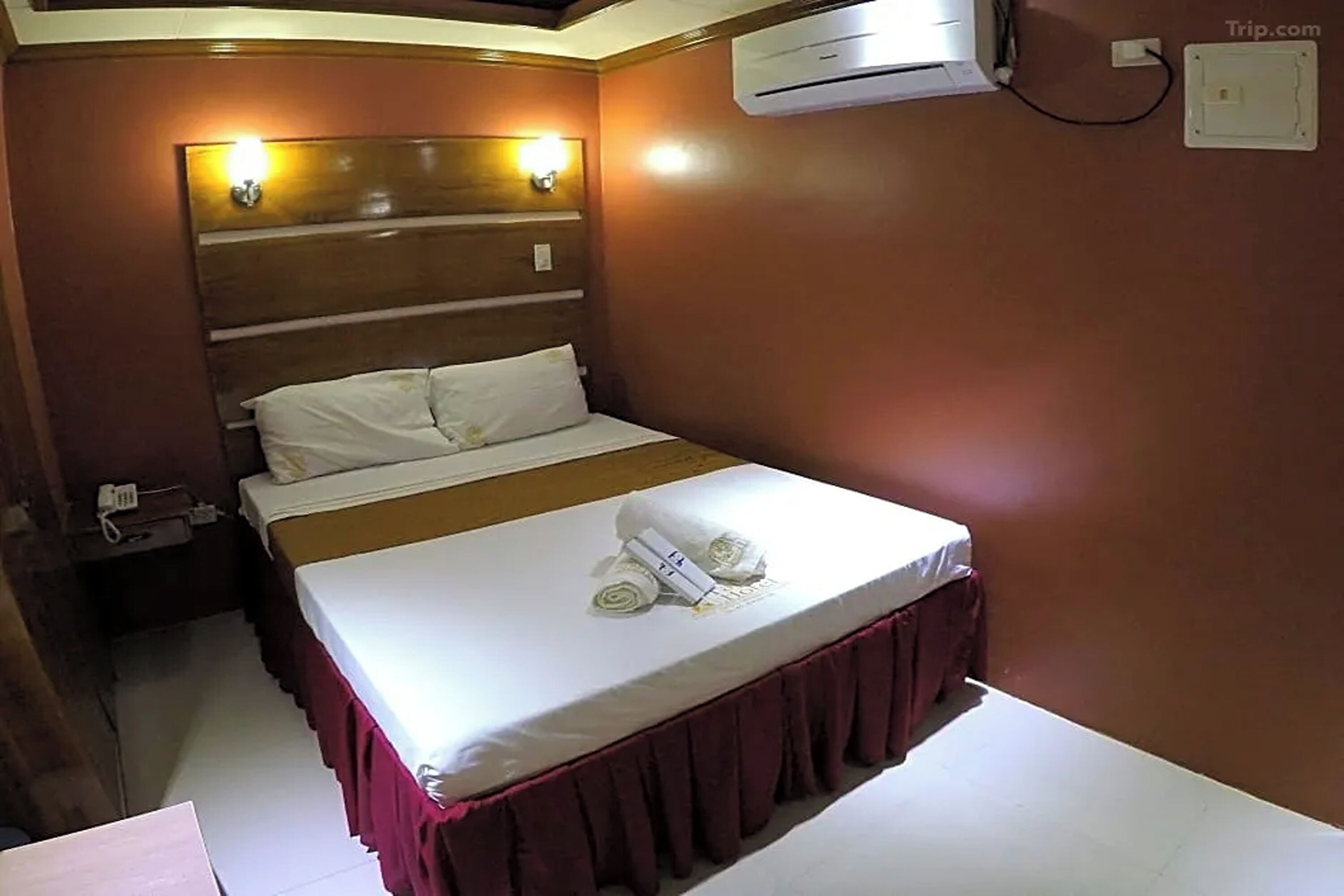 A hotel room with a bed, a nightstand, an air conditioning unit, and a wall-mounted light fixture.