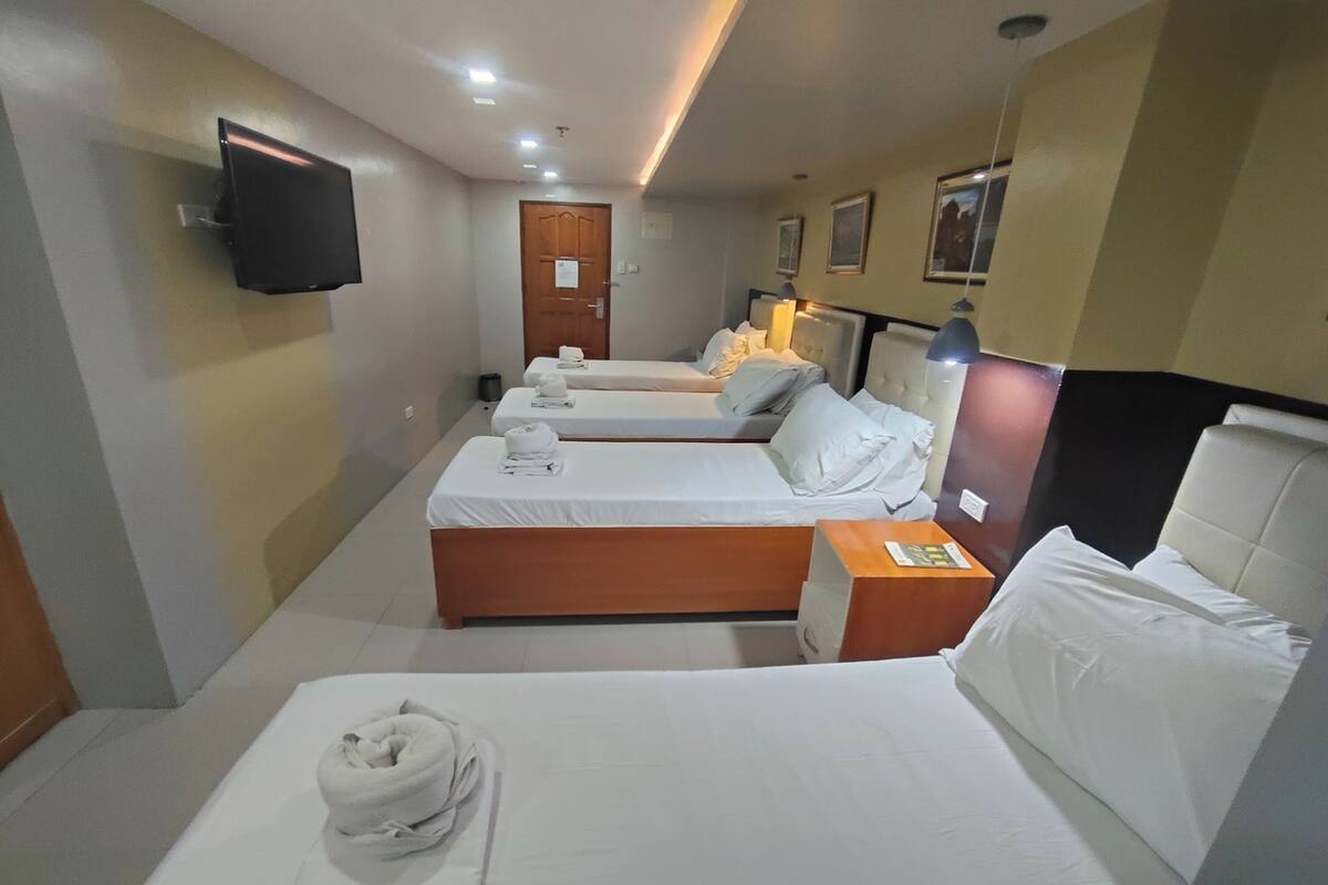 Une chambre d’hôtel avec trois lits simples, un téléviseur à écran plat fixé au mur, une table de chevet avec une lampe et une porte avec une pancarte.