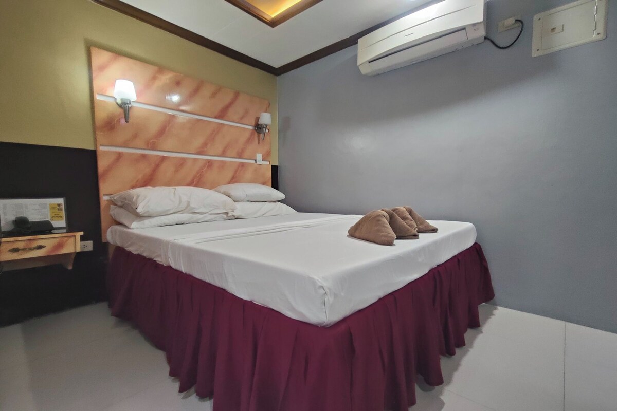 A neatly made bed with a white mattress and maroon skirt, a wooden headboard with light fixtures, and an air conditioning unit on the wall.