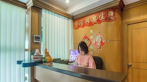 Un comptoir d’accueil avec une femme assise derrière, un mur décoratif orné de caractères chinois et une petite statue posée sur le comptoir.