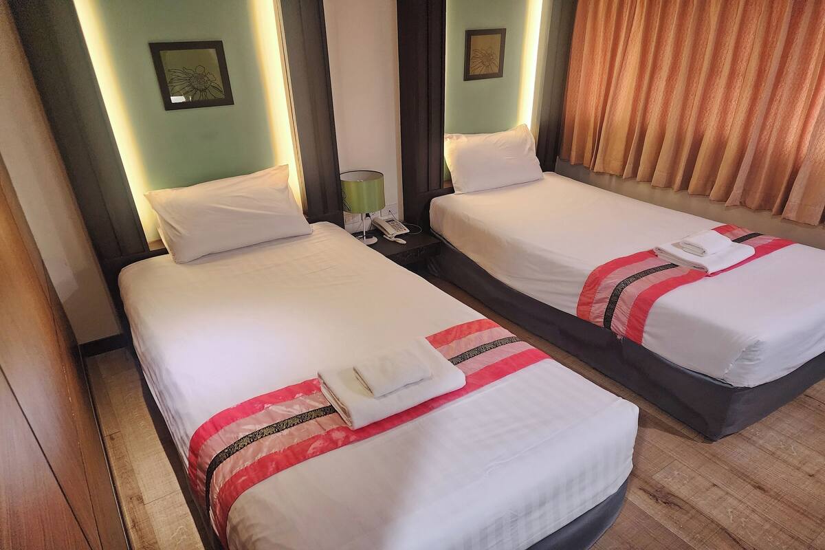 A hotel room with two single beds, each with a white comforter and a red-striped blanket. There is a bedside table with a lamp and a phone.