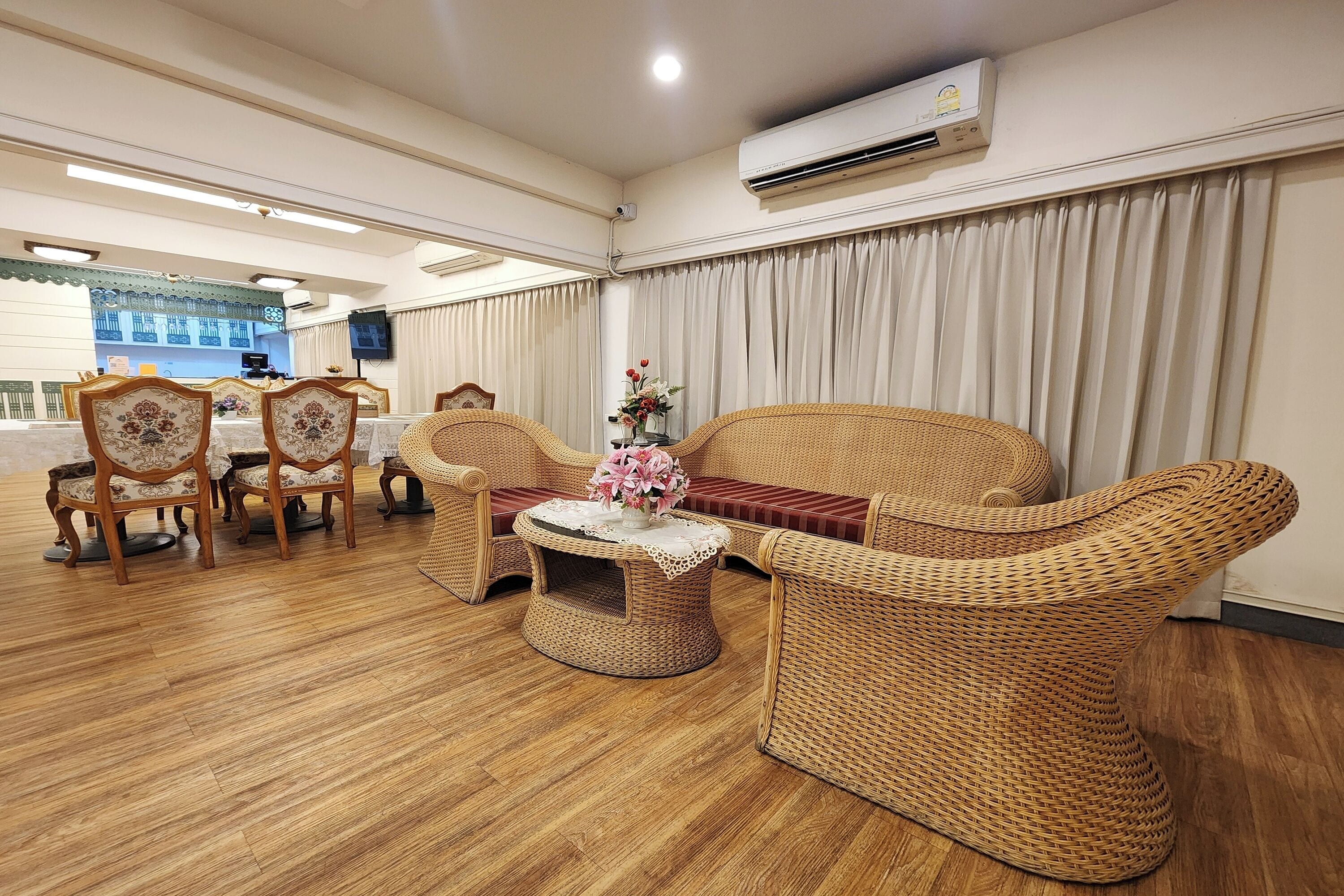 A room with a dining table, chairs, a wicker sofa, a small round table with a lace tablecloth, and an air conditioning unit on the wall.