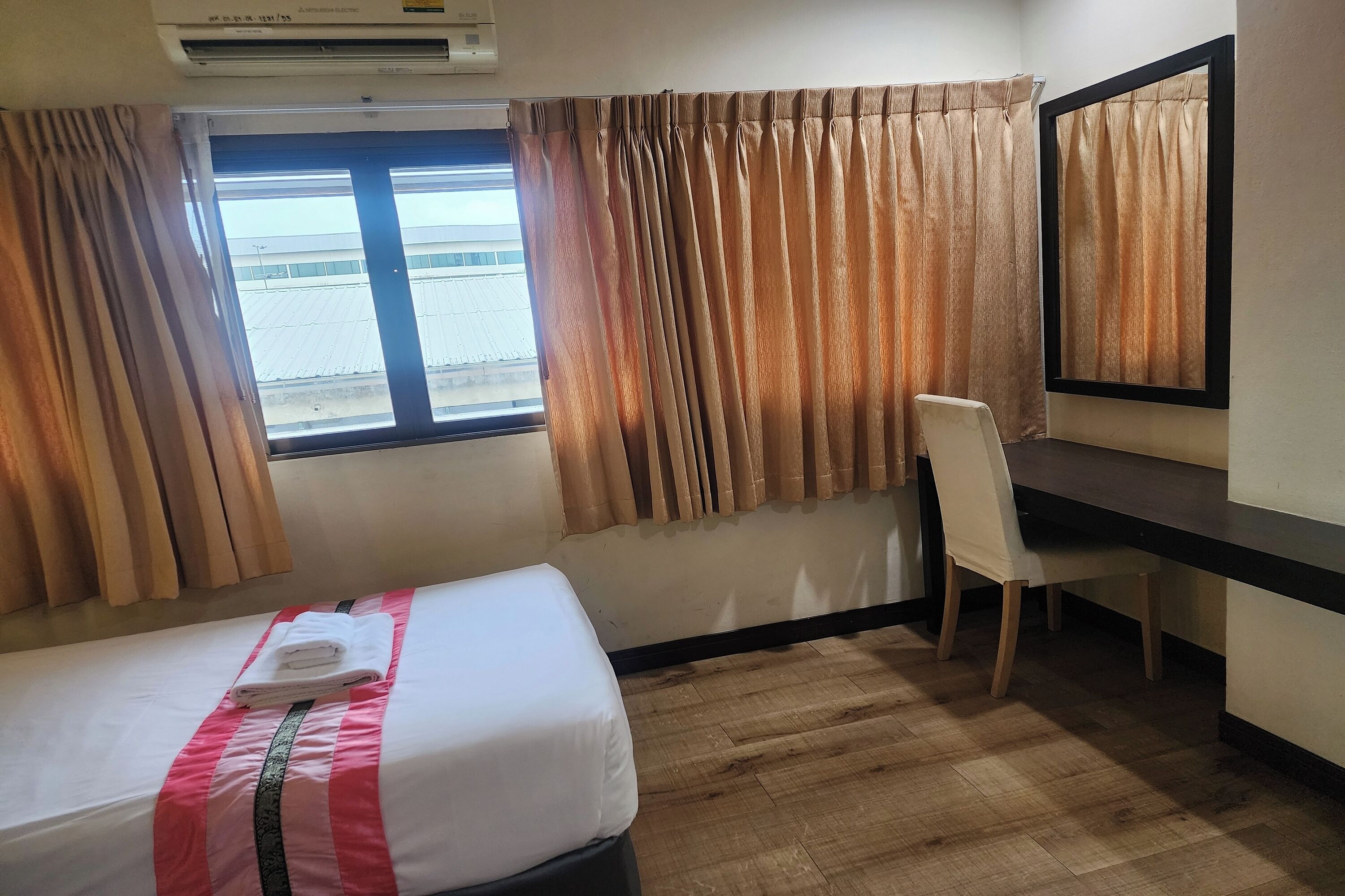 Quarto de hotel com cama, escrivaninha, cadeira, janela com cortinas e ar condicionado.