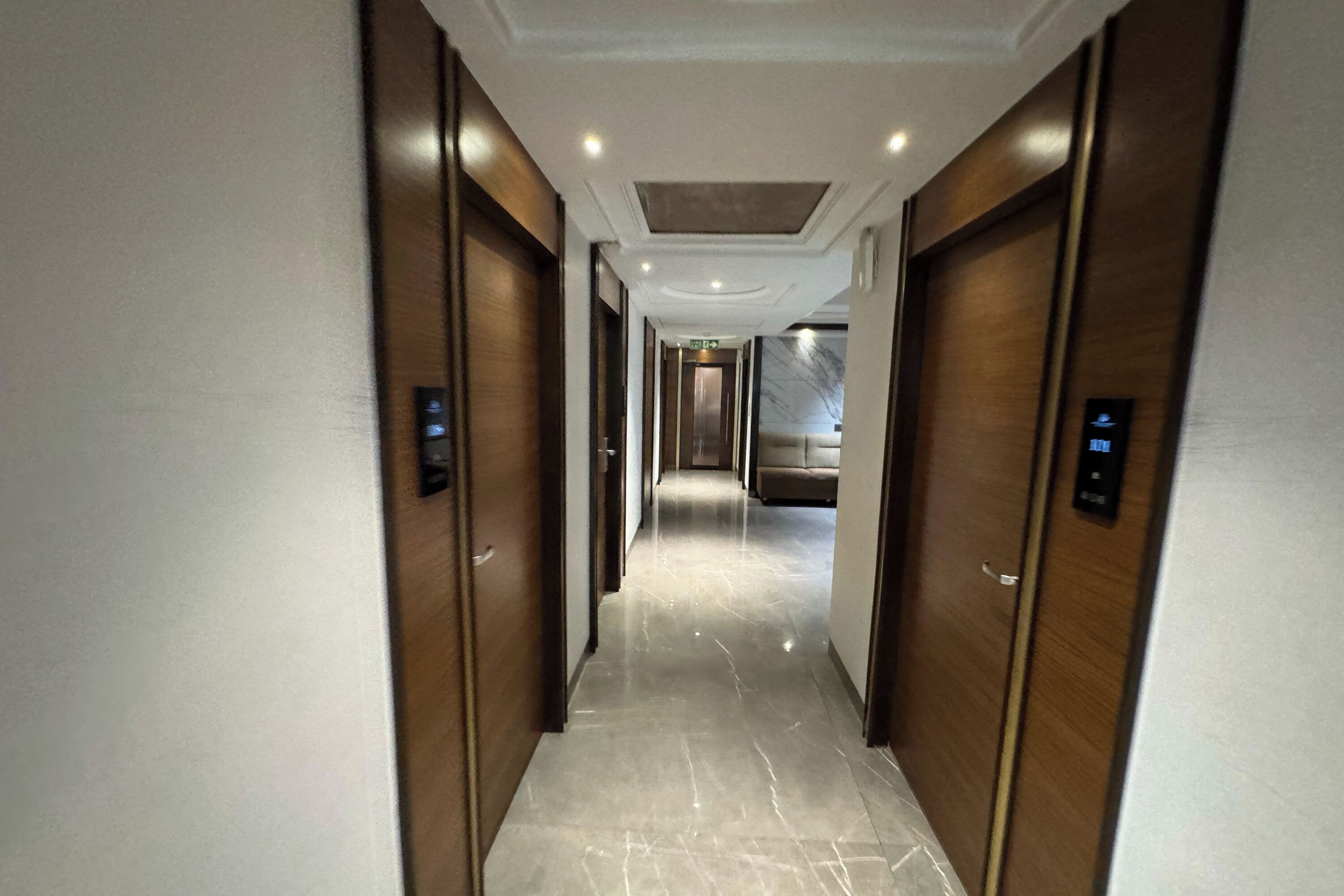 A modern hallway with marble flooring and wooden doors, leading to a room with a marble wall and a large mirror.