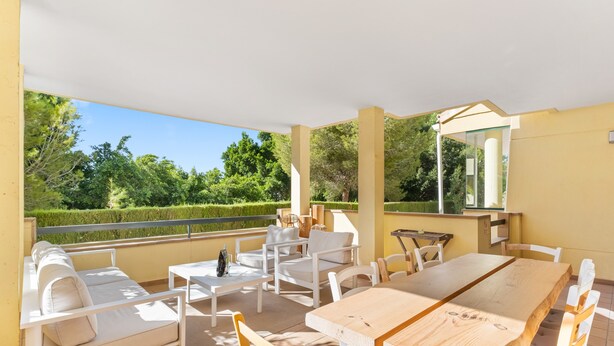 Une terrasse extérieure couverte, aménagée avec des meubles blancs, une table en bois et offrant une vue sur un jardin bien entretenu.