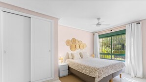 Une chambre à coucher comprenant un lit, une table de chevet, un ventilateur de plafond, une fenêtre avec des rideaux et une vue sur la verdure à l’extérieur.