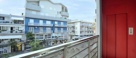 A balcony with a red door, overlooking a row of multi-story buildings with balconies, some with plants.