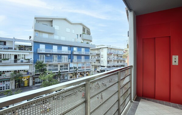 Un balcón con una puerta roja, con vistas a una hilera de edificios de varios pisos con balcones, algunos con plantas.