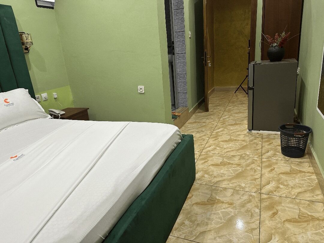 A hotel room with a bed, a small table with a phone, a wall-mounted lamp, a door, a refrigerator, and a trash bin.