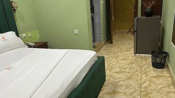 A hotel room with a bed, a small table with a phone, a wall-mounted lamp, a door, a refrigerator, and a trash bin.