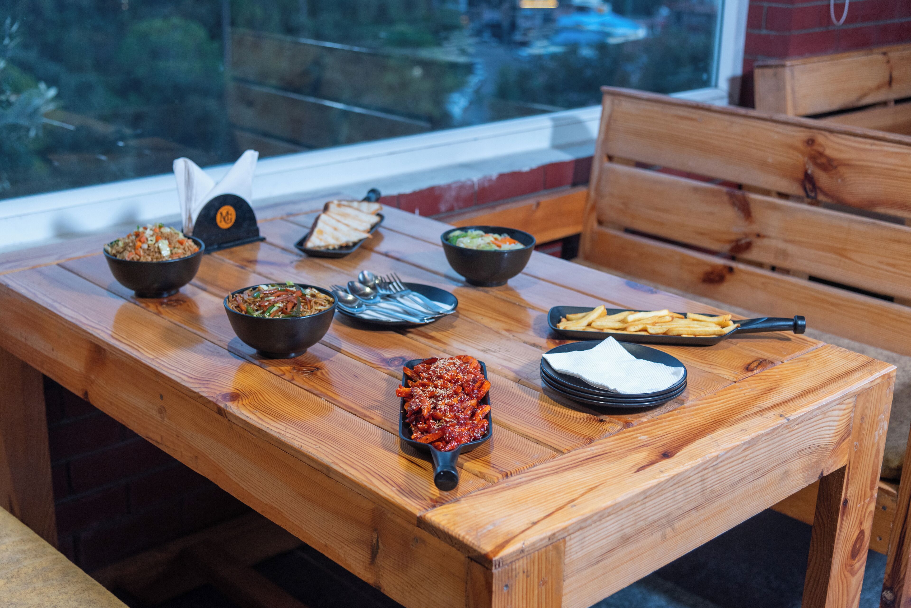 A wooden table with various dishes including a bowl of salad, a bowl of soup, a skillet with pasta, a plate of fries, and a small bowl of sauce.