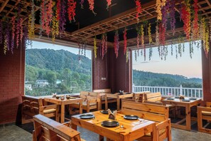 A restaurant with wooden tables and benches, hanging flower decorations, and a view of a forested hill.