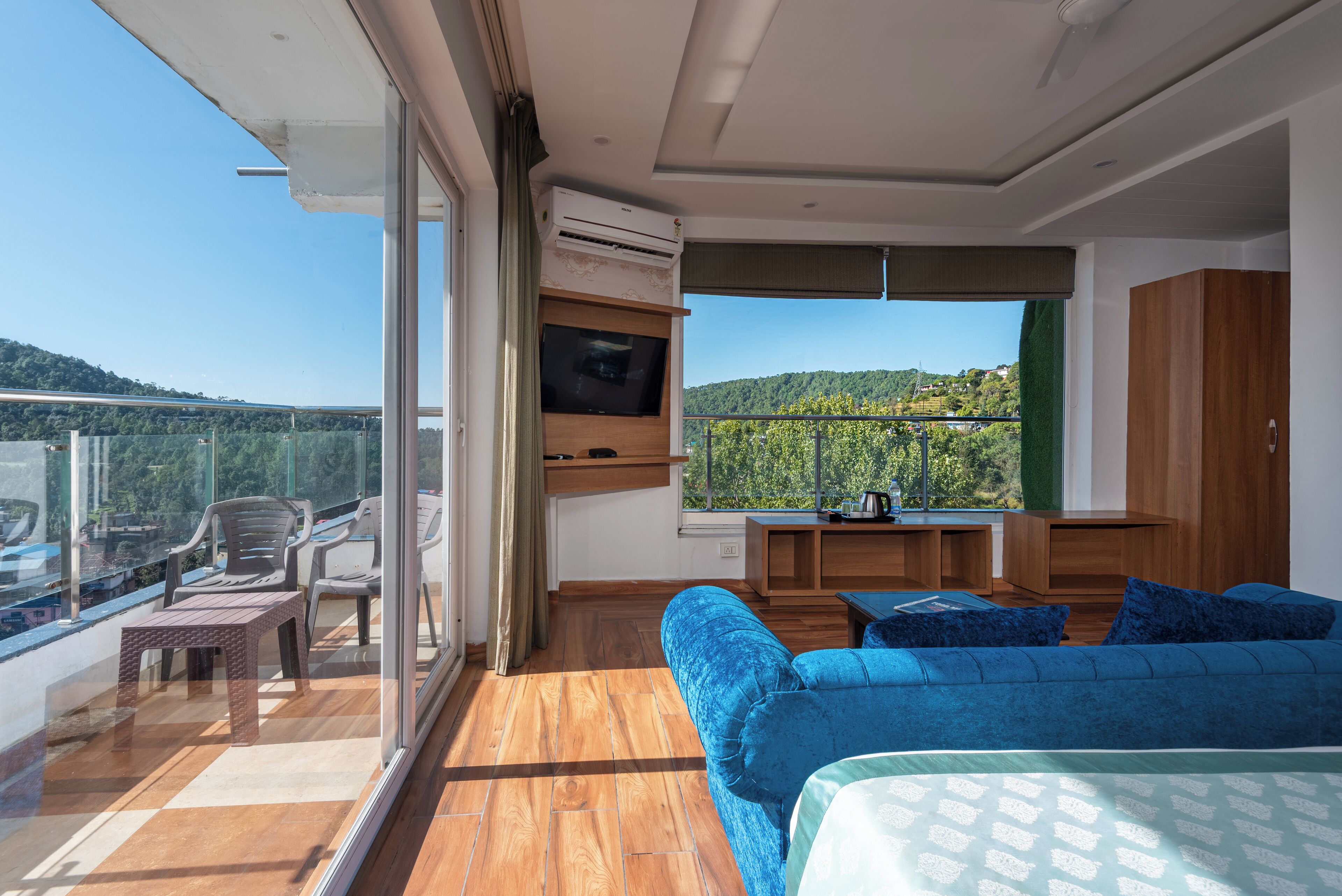 A balcony with a wooden floor, a blue sofa, a TV, and a view of a green landscape.