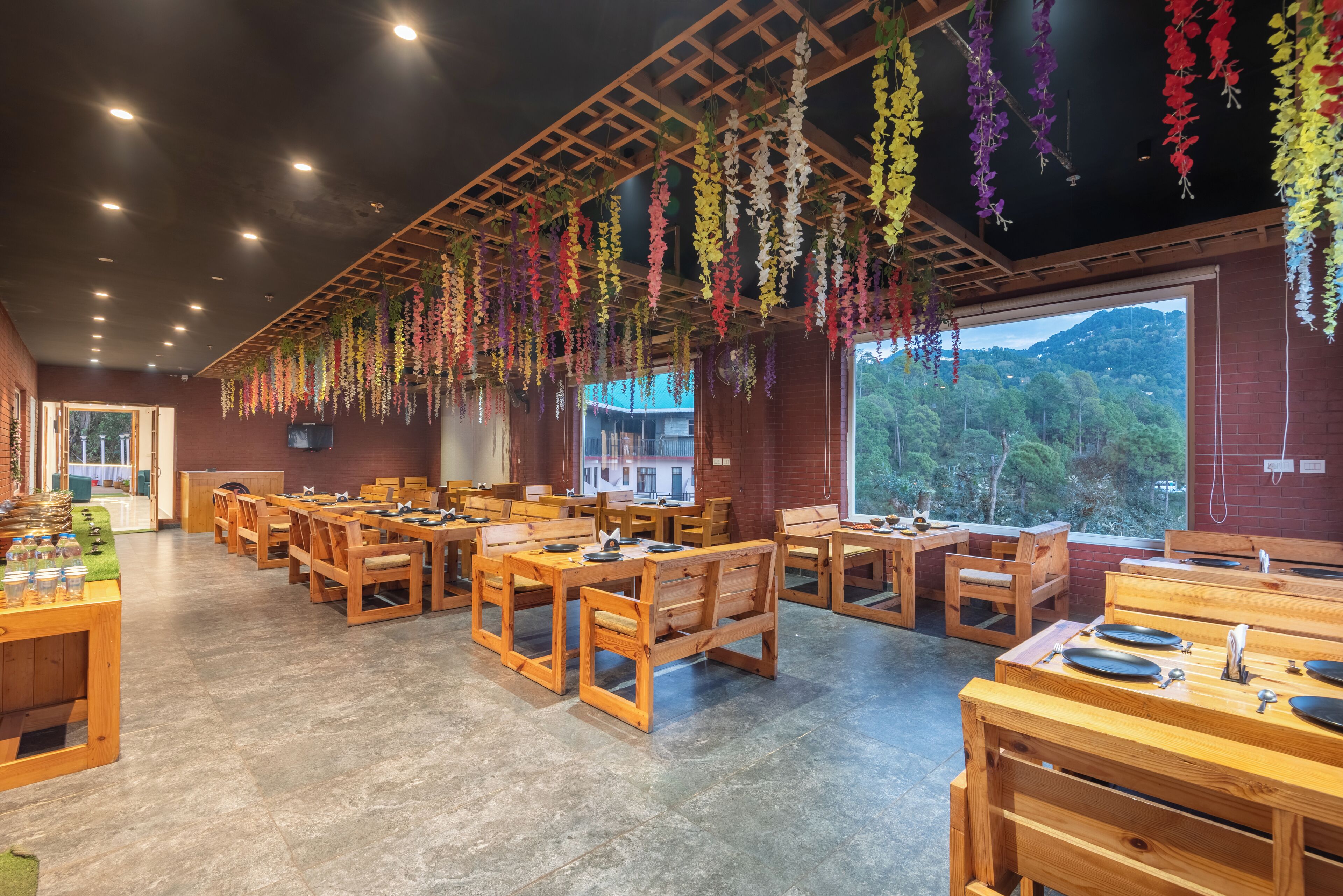 A restaurant with wooden tables and chairs, hanging flower decorations, and a view of a mountainous landscape through large windows.