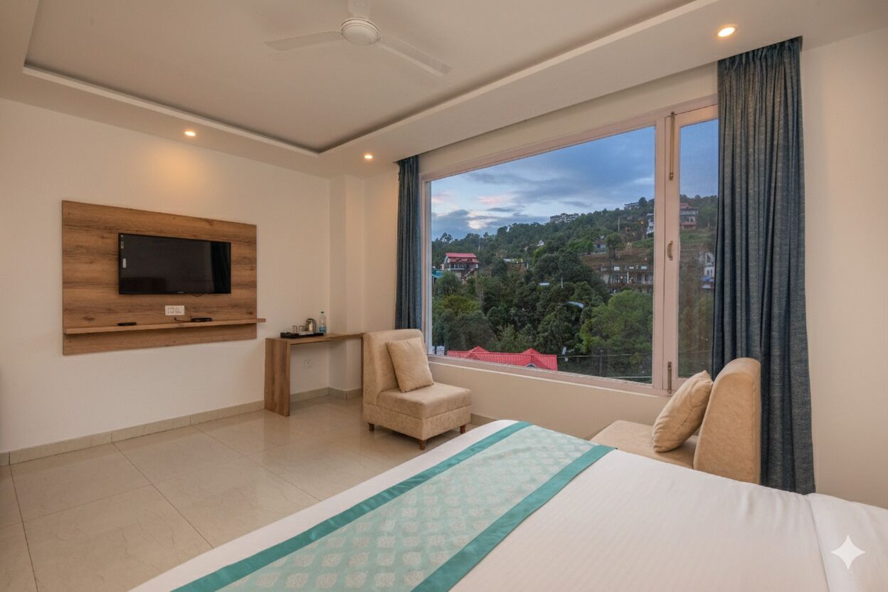 Habitació d'hotel amb un llit, un televisor fixat a la paret, una cadira, una taula petita i una finestra gran amb vista a un paisatge urbà.