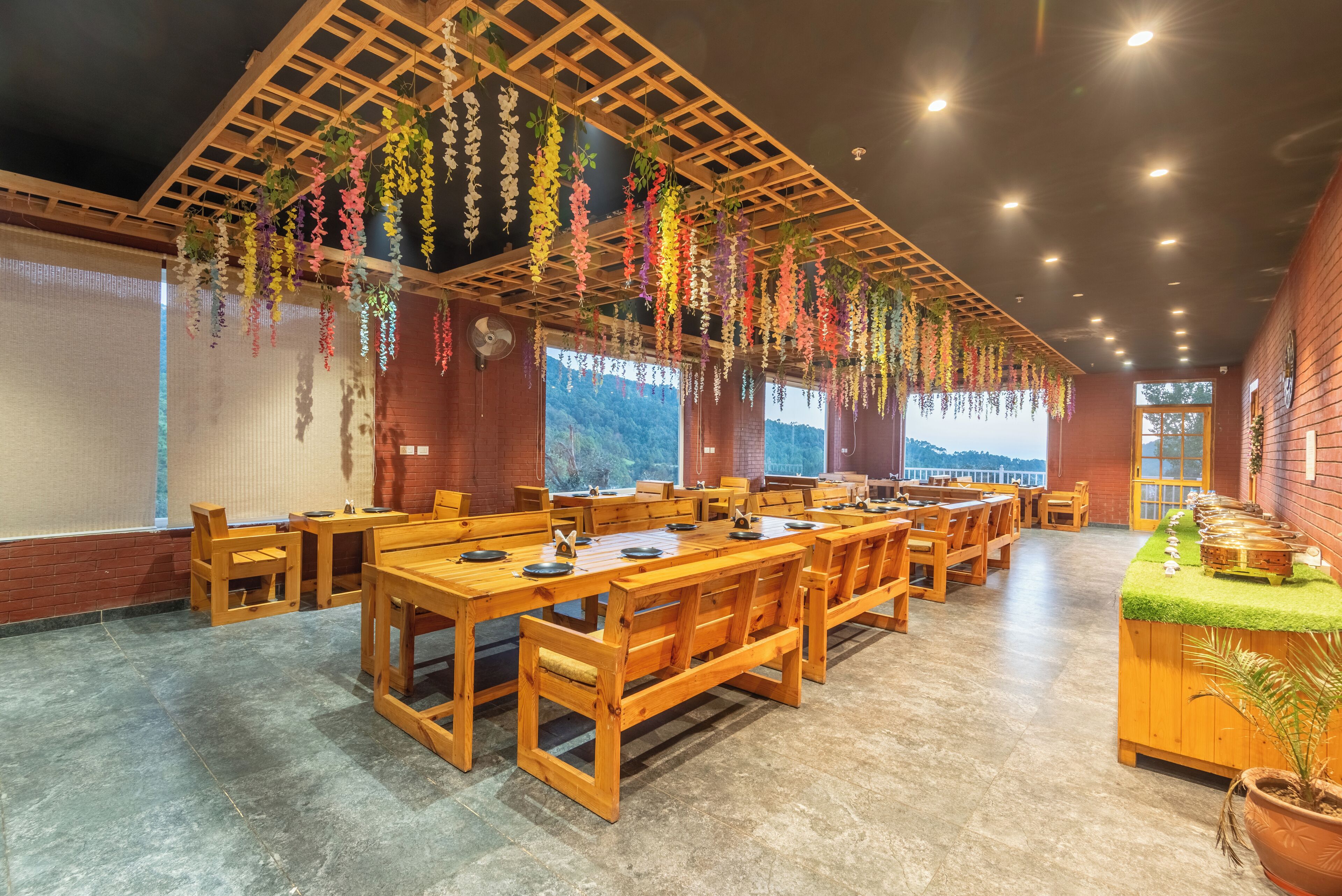 A restaurant with wooden tables and benches, hanging plants, and large windows offering a view of the outdoors.