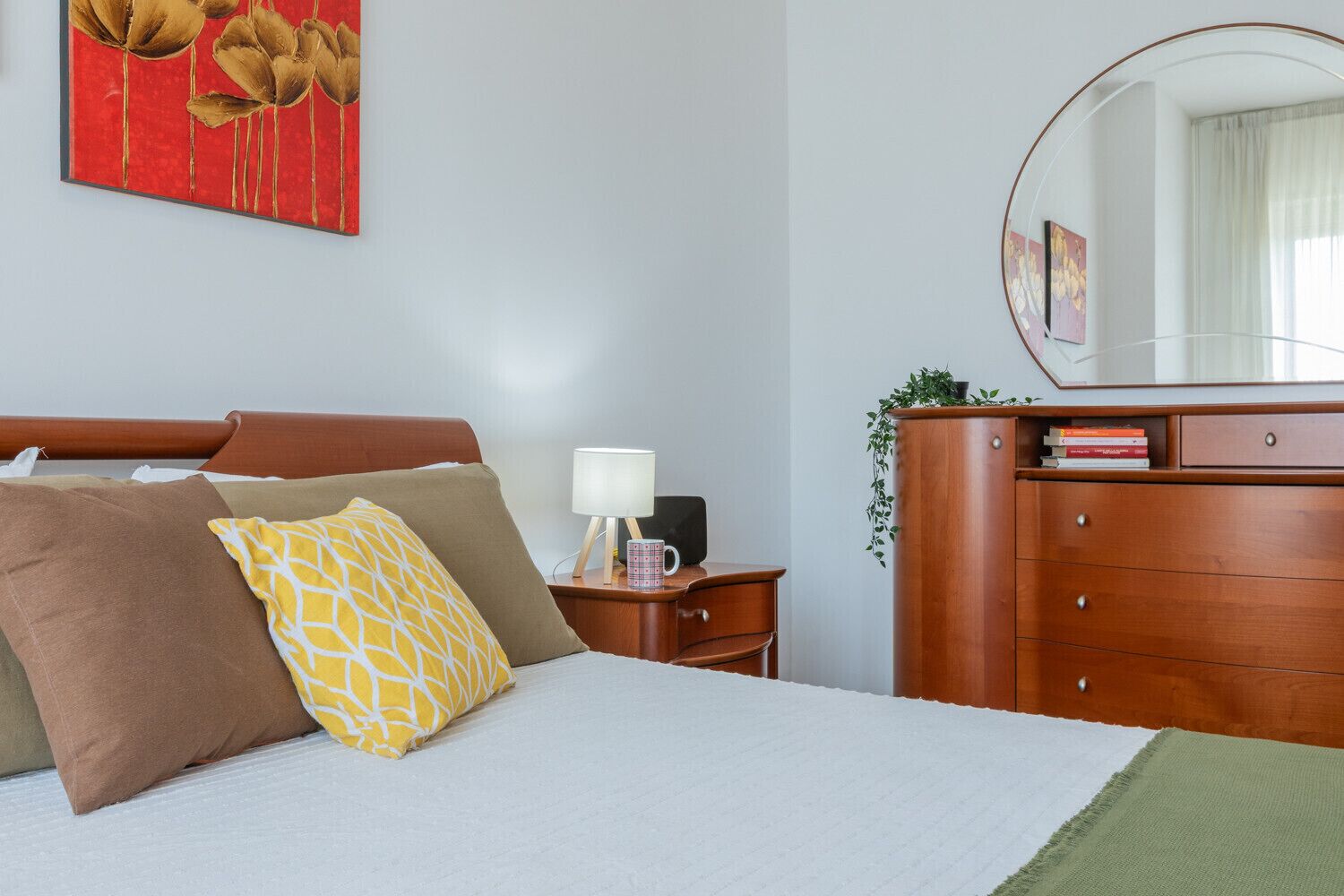 Ein ordentlich bezogenes Bett mit Kissen, ein Nachttisch mit Lampe, eine Kommode mit Spiegel und ein rotes Blumenbild an der Wand.