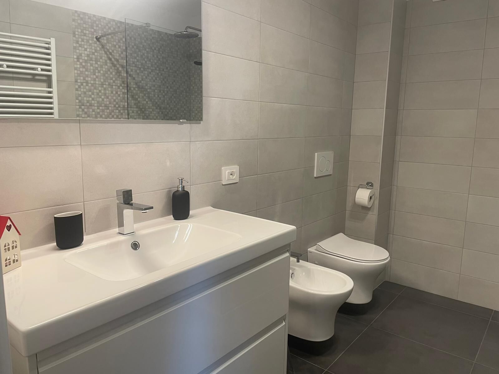 Une salle de bain moderne avec un lavabo blanc, deux toilettes, une douche et un miroir.