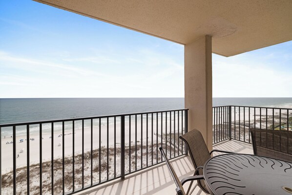 A balcony with a view of the beach, a table, and chairs.