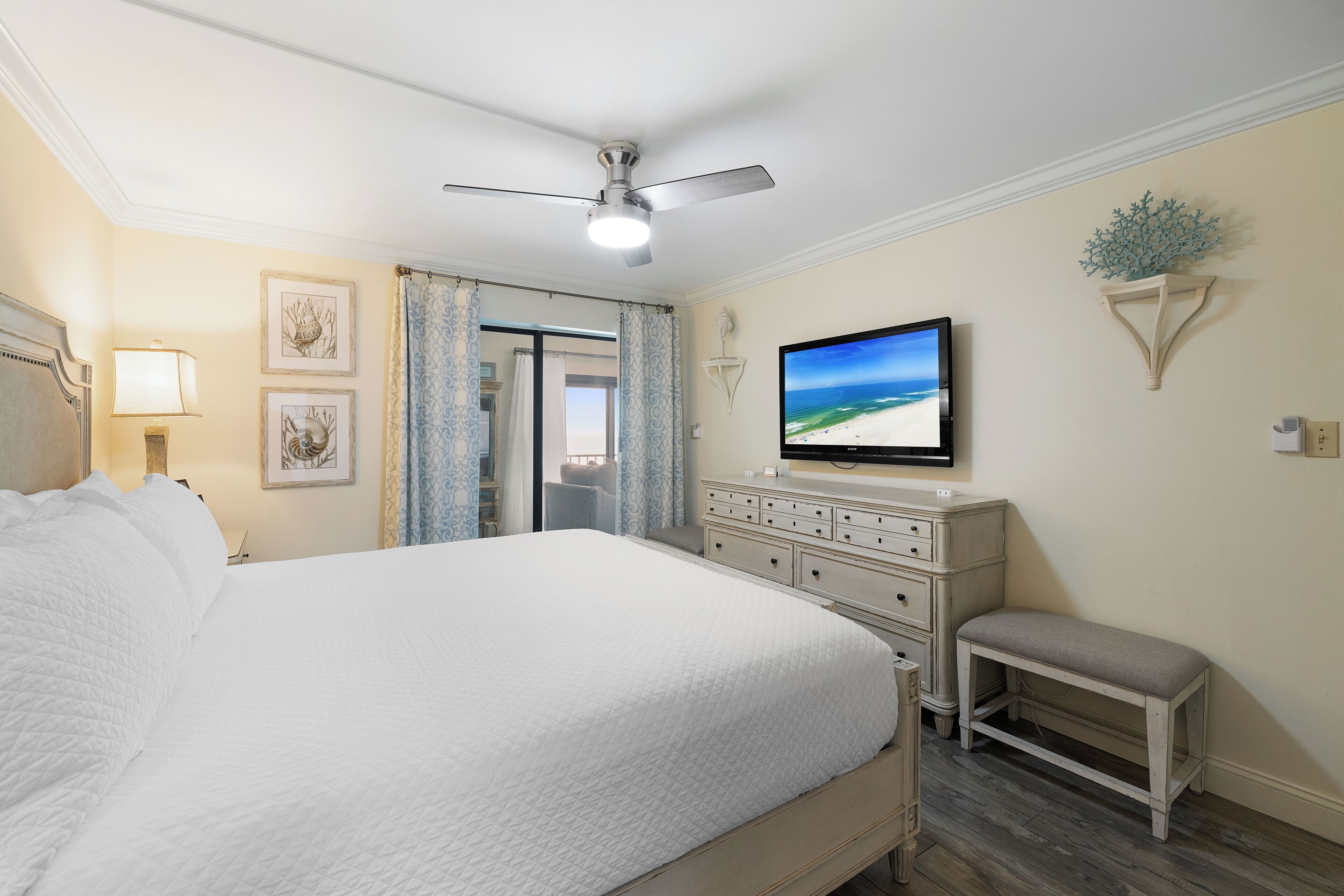 Une chambre bien rangée, avec un grand lit, un ventilateur de plafond, une télévision et un petit banc.