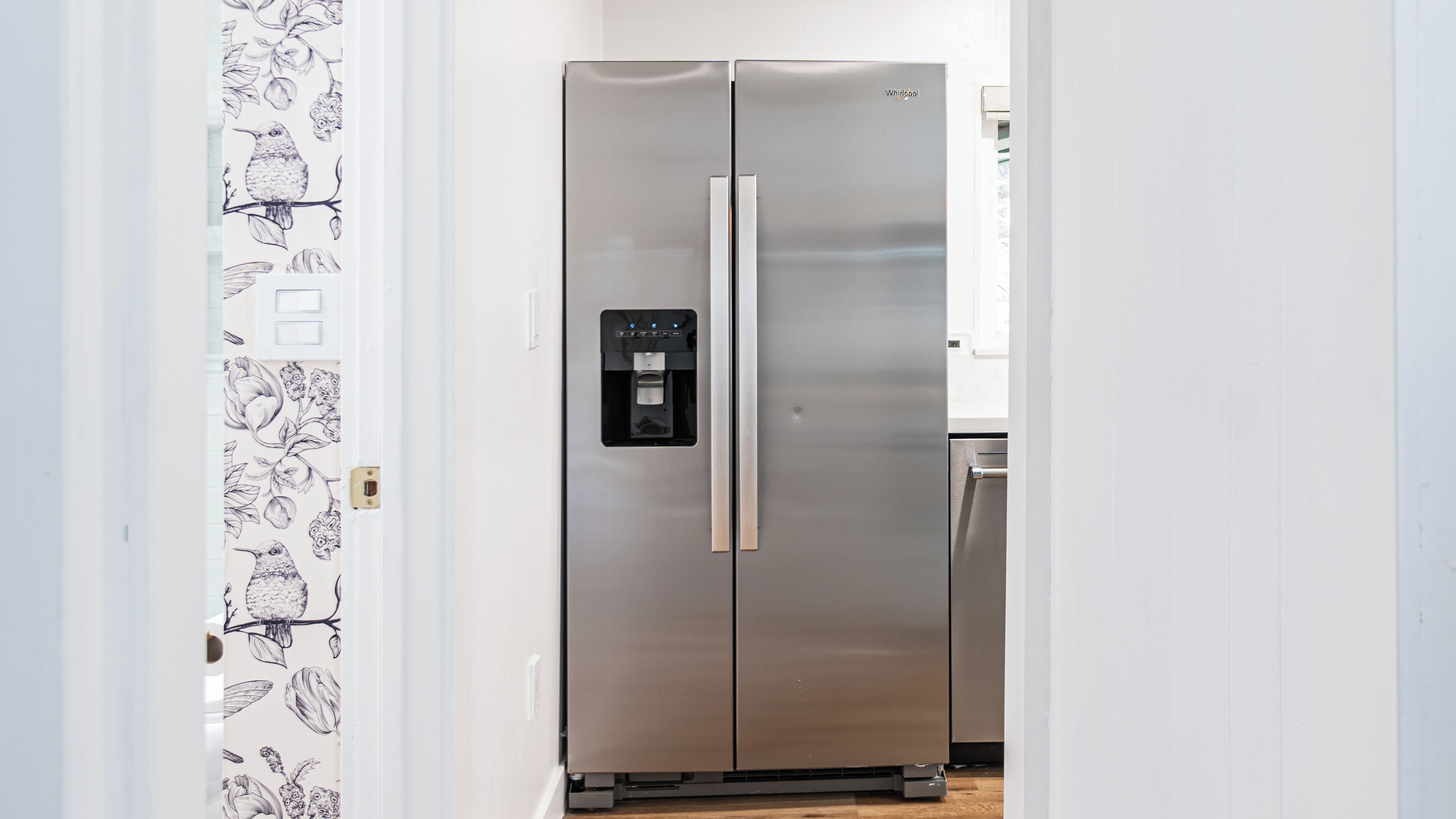 Un refrigerador Whirlpool de acero inoxidable con dispensador de agua, situado en un pasillo con puerta blanca y papel tapiz floral.