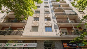 Un edificio de varios pisos con balcones, locales comerciales abajo y un árbol en primer plano.