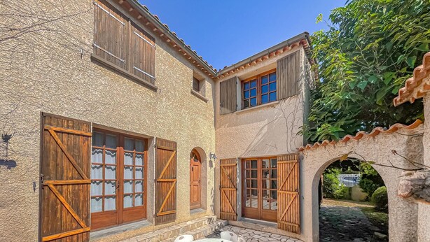 Un edificio de piedra con contraventanas de madera, un patio con mesa y sillas, y un arco que da a un jardín.