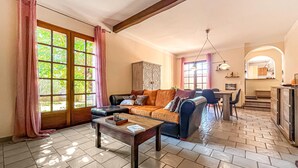 Amplia sala de estar con sofá, una mesa de centro y una mesa de comedor. Se ve una ventana grande con cortinas, un candelabro y una cocina al fondo.
