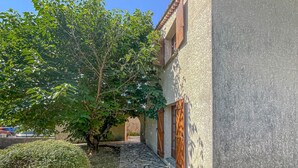 Un estrecho sendero de piedra que conduce a un edificio con fachada rústica, contraventanas de madera y un árbol que da sombra.