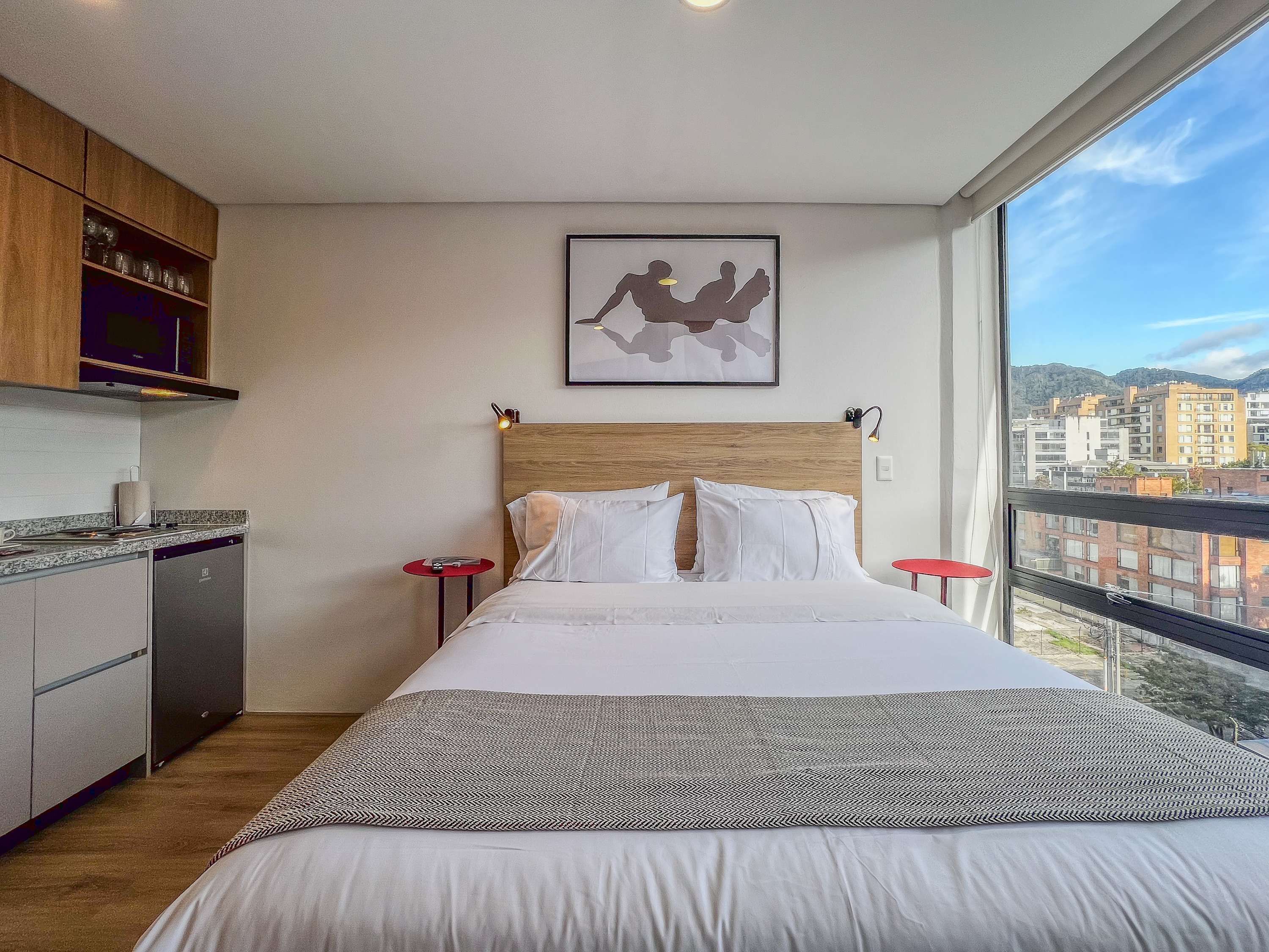 A hotel room with a bed, a small kitchen area, and a large window offering a city view.
