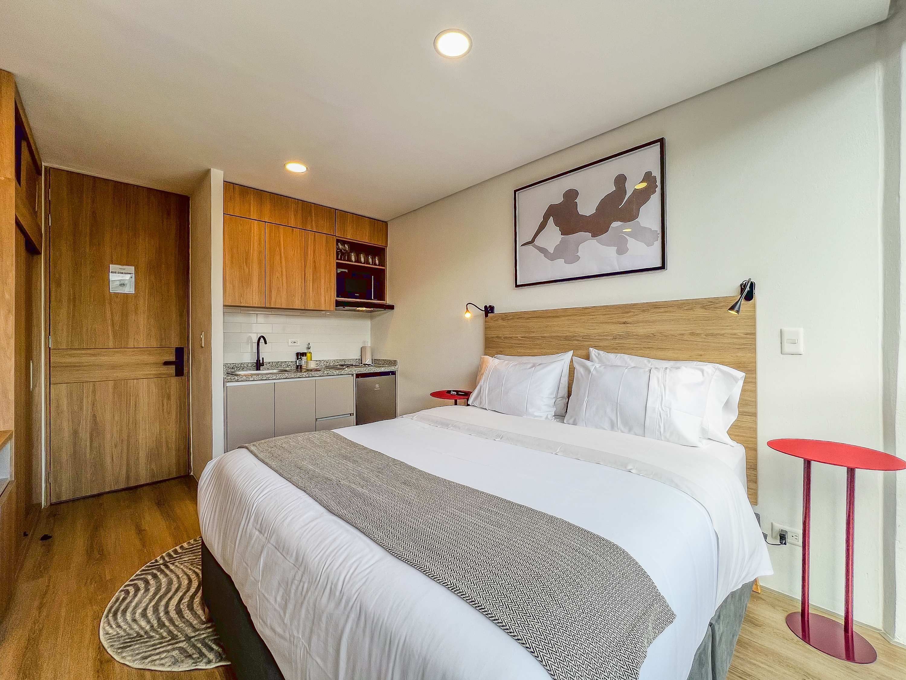 A hotel room with a bed, a small kitchenette, a wooden door, and a framed picture on the wall.