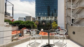 Eine Dachterrasse mit Tisch und zwei Stühlen, mit Blick auf eine Stadtlandschaft aus modernen Gebäuden und Grünflächen.