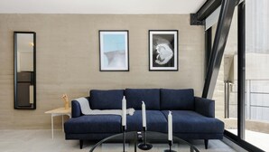 Ein modernes Wohnzimmer mit einem blauen Sofa, zwei gerahmten Kunstwerken, einem runden Glastisch mit Kerzen und einem schlichten Beistelltisch.