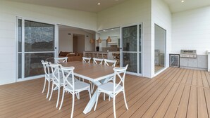 Une vaste terrasse dotée d’une table en bois et de chaises blanches, un coin cuisine avec une cuisinière, et des portes-fenêtres coulissantes donnant sur un espace intérieur.