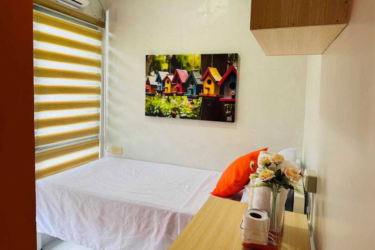 Een slaapkamer met een bed, een aan de muur hangend beeld van kleurrijke vogelhuisjes, een raam met jaloezieën en een nachtkastje met een bloevas.
