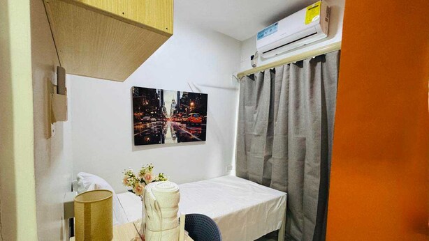 Een kleine hotelkamer met een bed, een bureau met een lamp, een airconditioner en een stadsgezichtschilderij aan de muur.