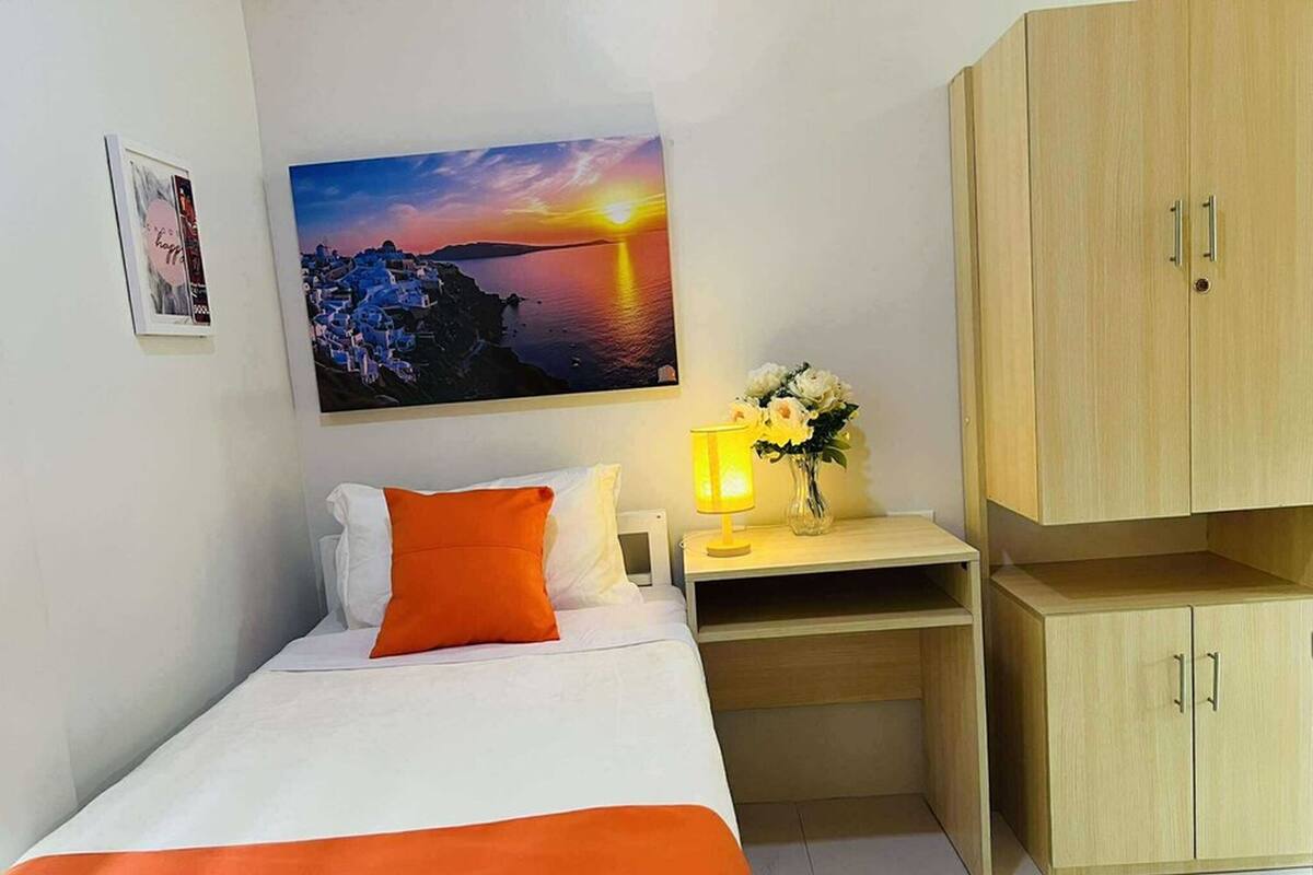 Een slaapkamer met een bed, nachtkastje, lamp, een vaas met bloemen en een houten garderobe.
